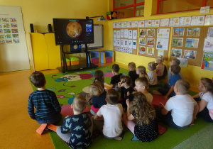 Grupa dzieci siedzi przed ekranem i z zaciekawieniem ogląda poszczególne planety.